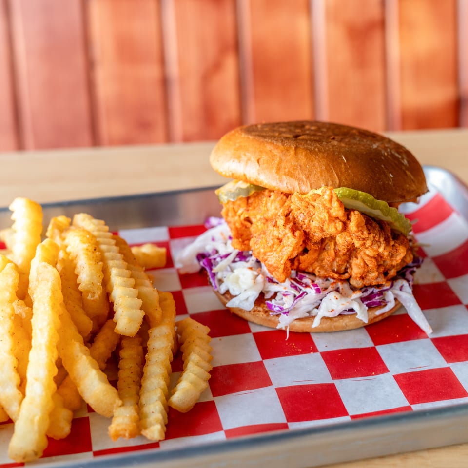 Spicy fried chicken sandwich with coleslaw and pickles, served with crinkle-cut fries on a metal tray.