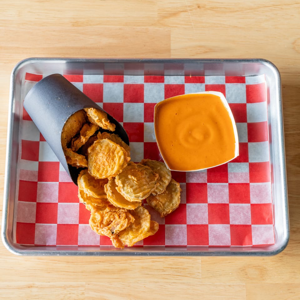 Fried pickles spilling from a black paper cone, with a side of orange dipping sauce on a metal tray.