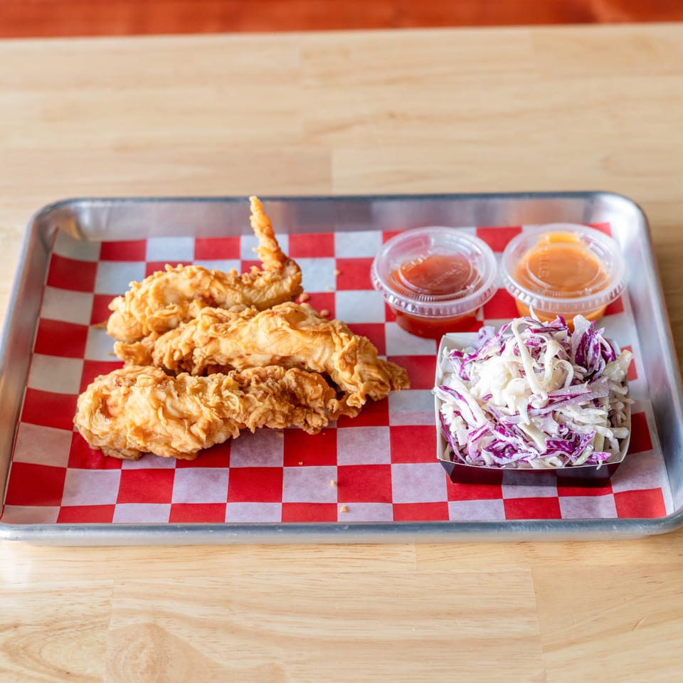 Crispy fried chicken tenders, coleslaw, and two dipping sauces on a metal tray with checkered paper.