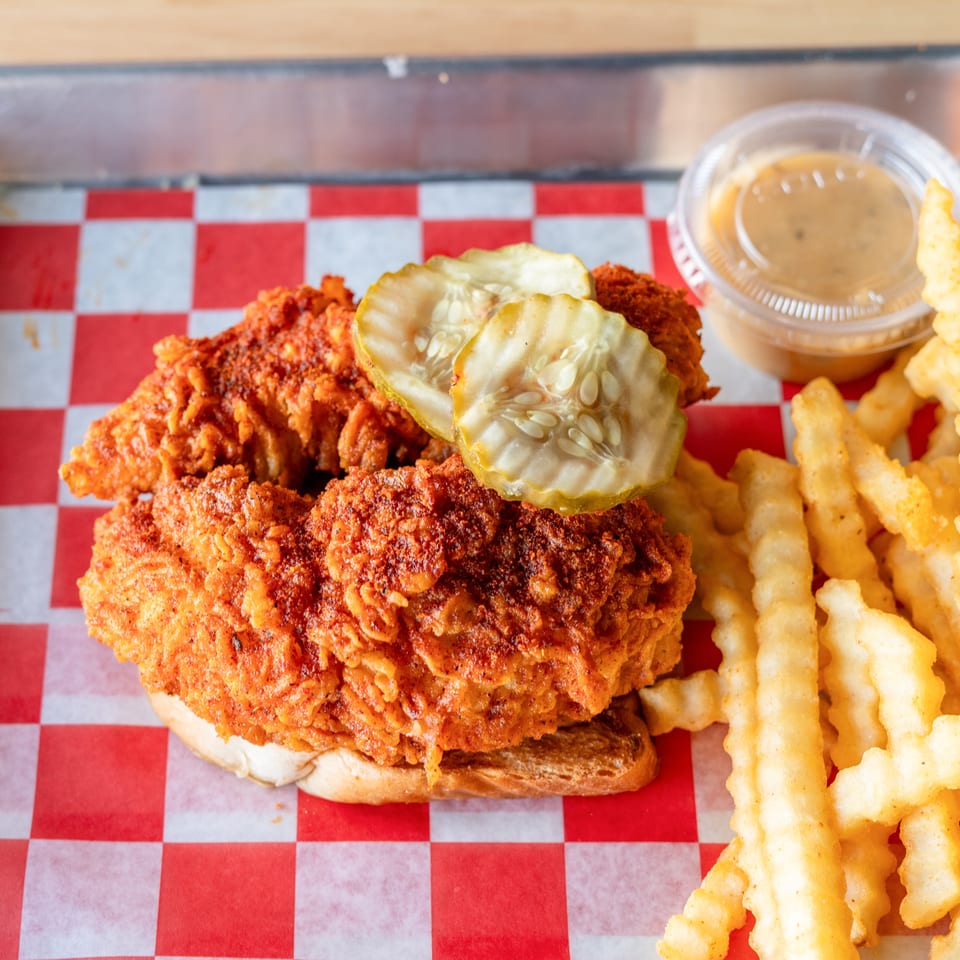 Spicy fried chicken sandwich with pickles and crinkle-cut fries on red and white checkered paper.