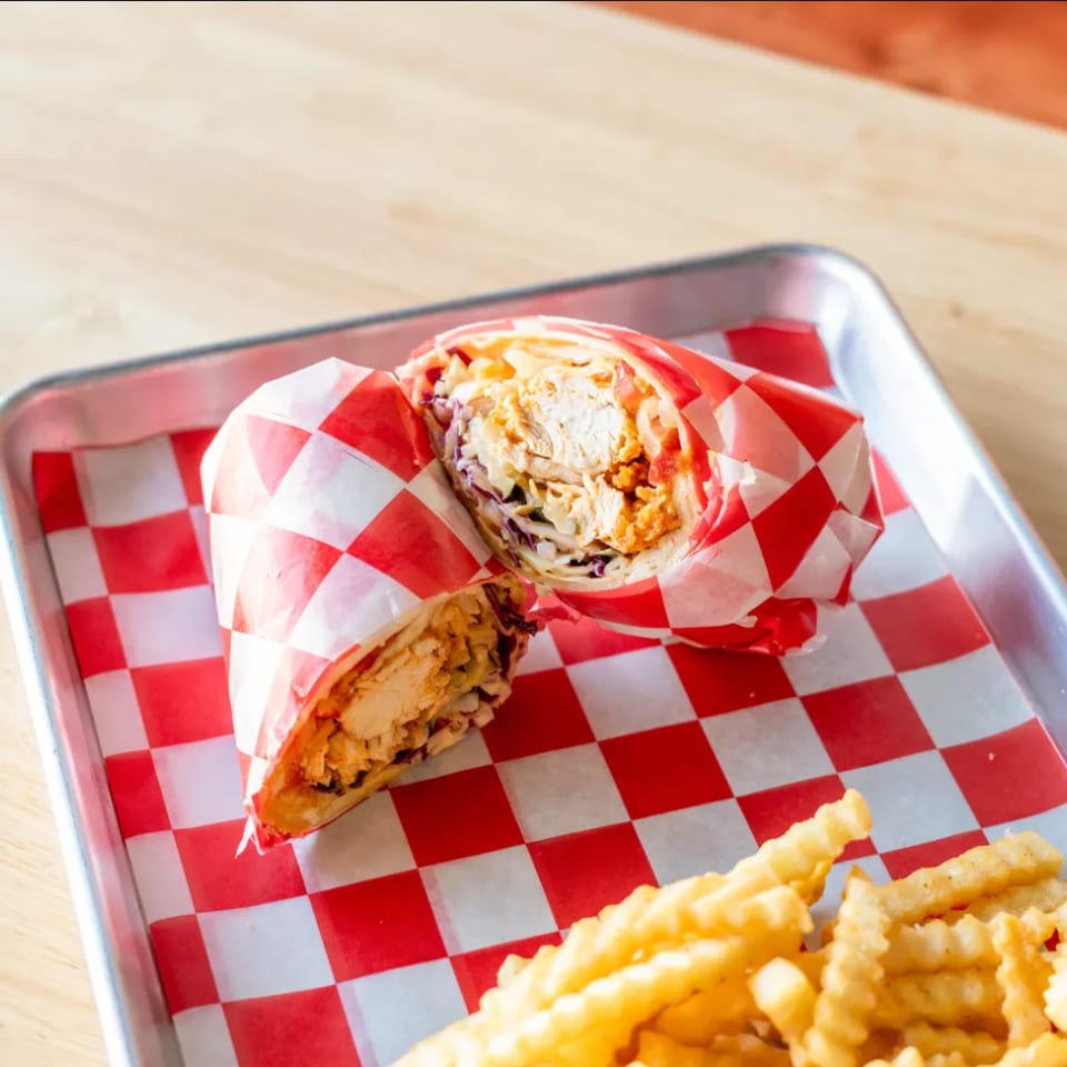A chicken wrap cut in half, wrapped in red and white checkered paper, with crinkle-cut fries.